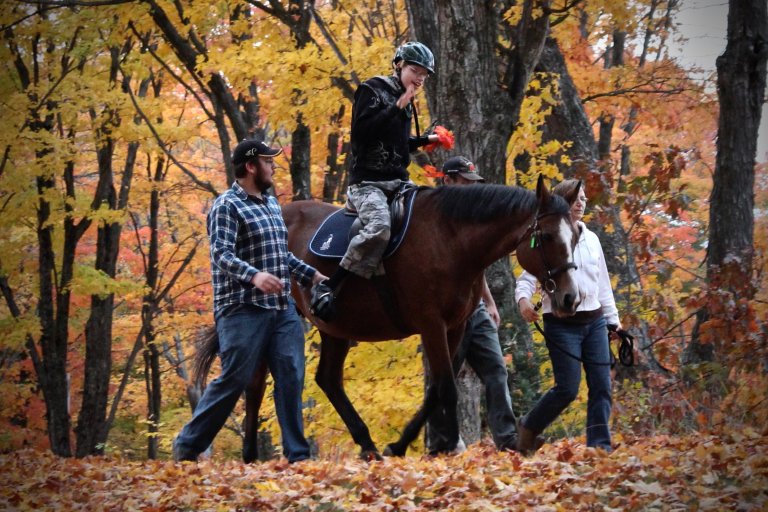 Boy riding a horse, while two volunteers are on the sides, helping to lead the horse through the forest