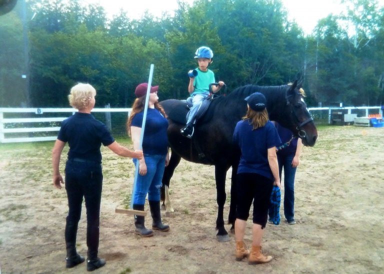 Four people in this image, one young boy on a horse, and three people around the boy and horse. One holding a stick and the other two standing beside the horse.