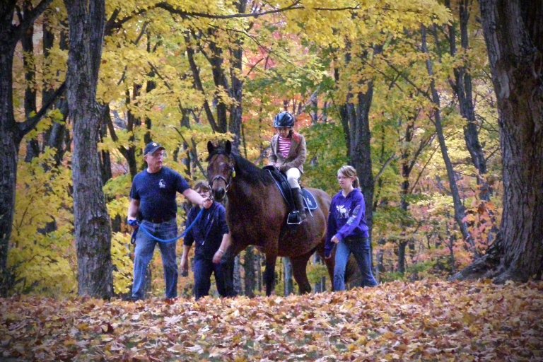 young girl riding a horse, while two volunteers are on the sides and an instructor is leading the horse through the forest