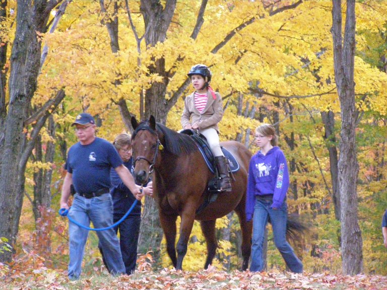 young girl riding a horse, while two volunteers are on the sides and an instructor is leading the horse through the forest